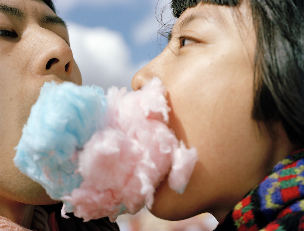A man and woman (Moro and Pixy) face each other. We see their faces in profile, close-up. Pink candyfloss emerges from Pixy's mouth, blue candyfloss from Moro's; it intermingles.