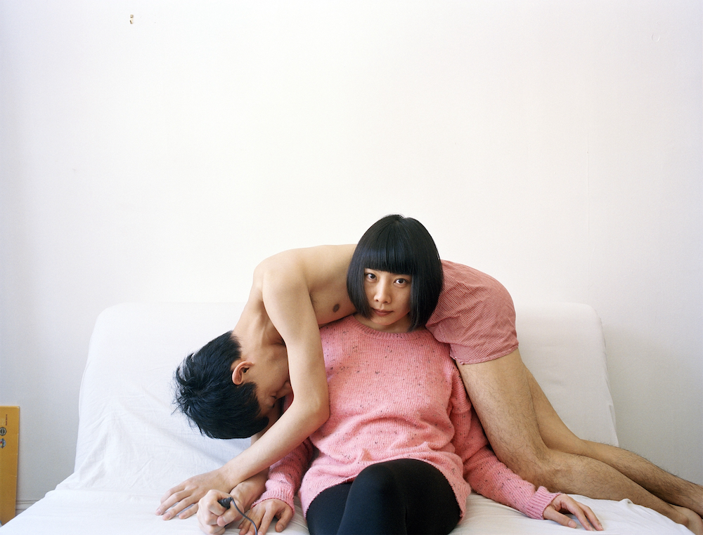 Pixy sits on a white sofa, facing the camera, wearing a pink jumper and black leggings. Moro is draped across her shoulders, wearing pink boxer shorts. His head is down and his eyes are closed. He holds the camera's shutter release in his hand.