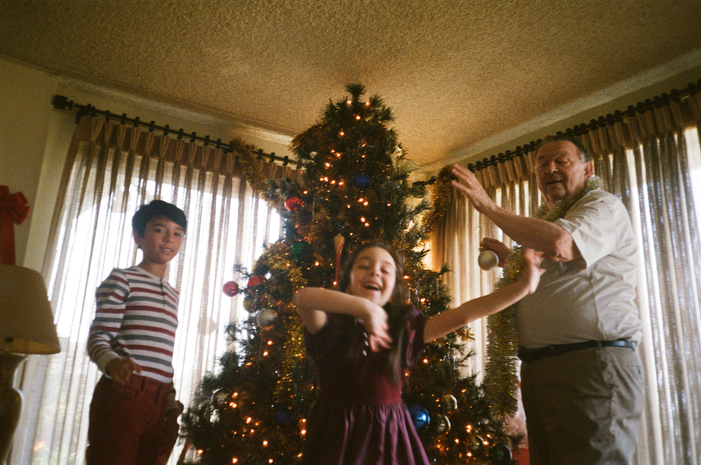 Colour photograph with grain texture. Two children (a boy and a girl) stand in front of a Christmas tree with an older man, who holds tinsel and baubles.