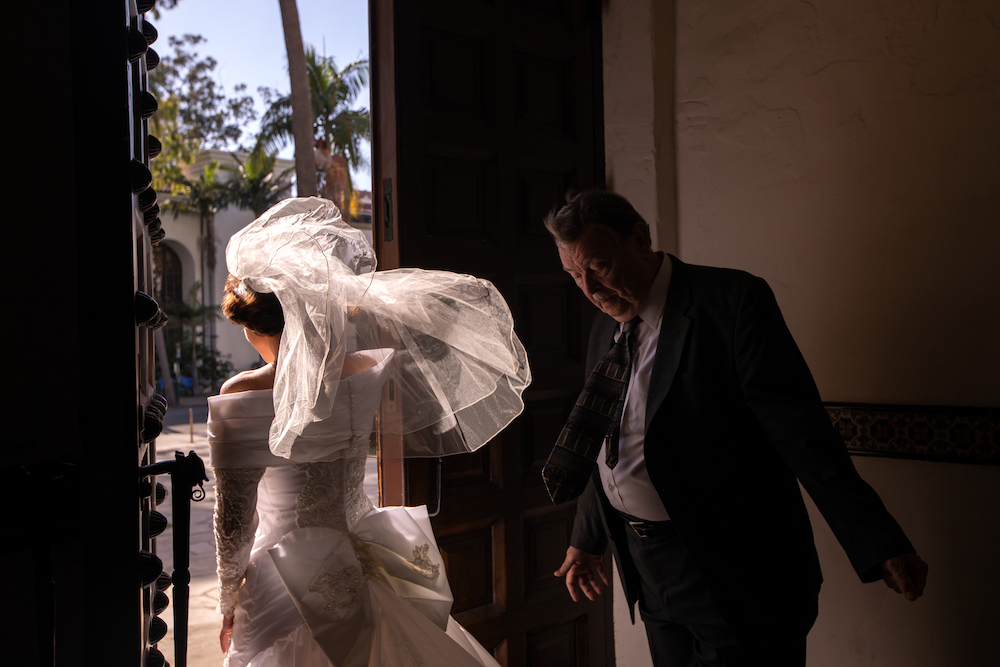 A woman and man are leaving a darkened corridor, stepping out into a sunny day. The woman faces away from the camera as she exits, her decorative wedding dress and veil billowing behind her. The man follows her, dressed in a suit and tie.