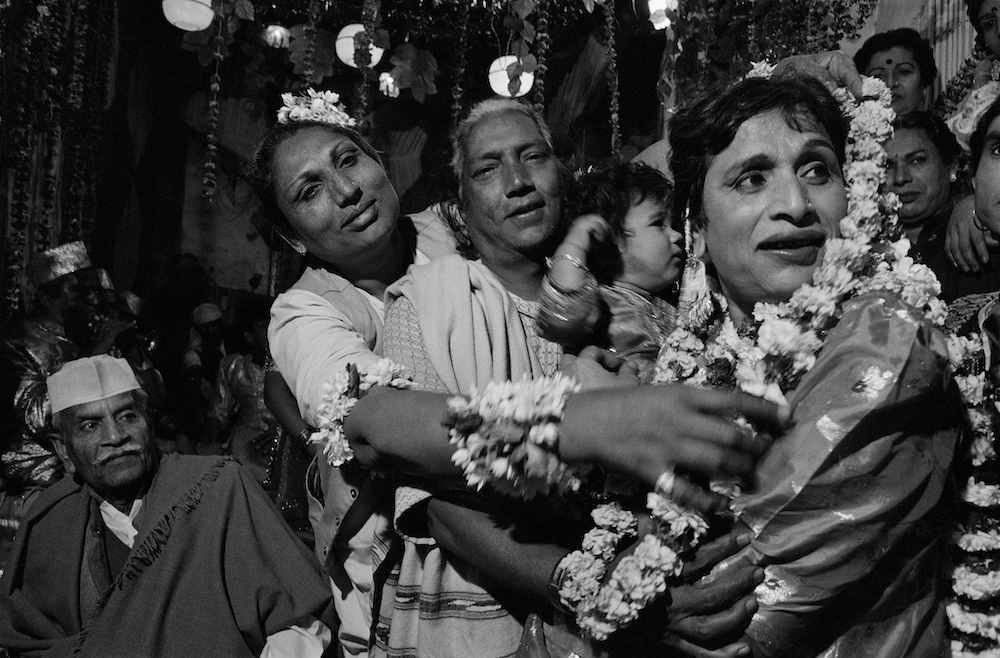 Black and white photograph. Three people and a small child are in the foreground, in front of a crowd of others. Mona Ahmed (front, right) greets guests by handing them garlands of flowers.