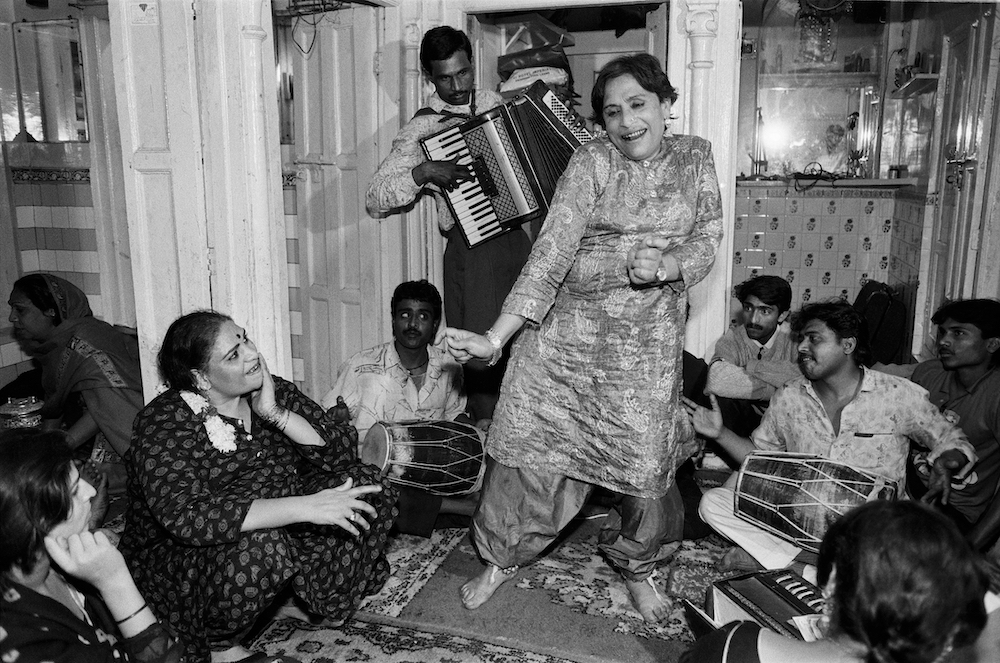 Black and white photograph. In an interior setting, Mona Ahmed dances. Gathered close by are musicians and onlookers, who sit in a circle around Mona. An accordion player stands in the background.