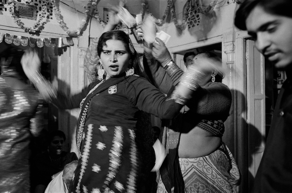 Black and white photograph. Blurred figures move across the scene, dancing at a party. More in focus, we see Shalu dancing and facing towards the camera.