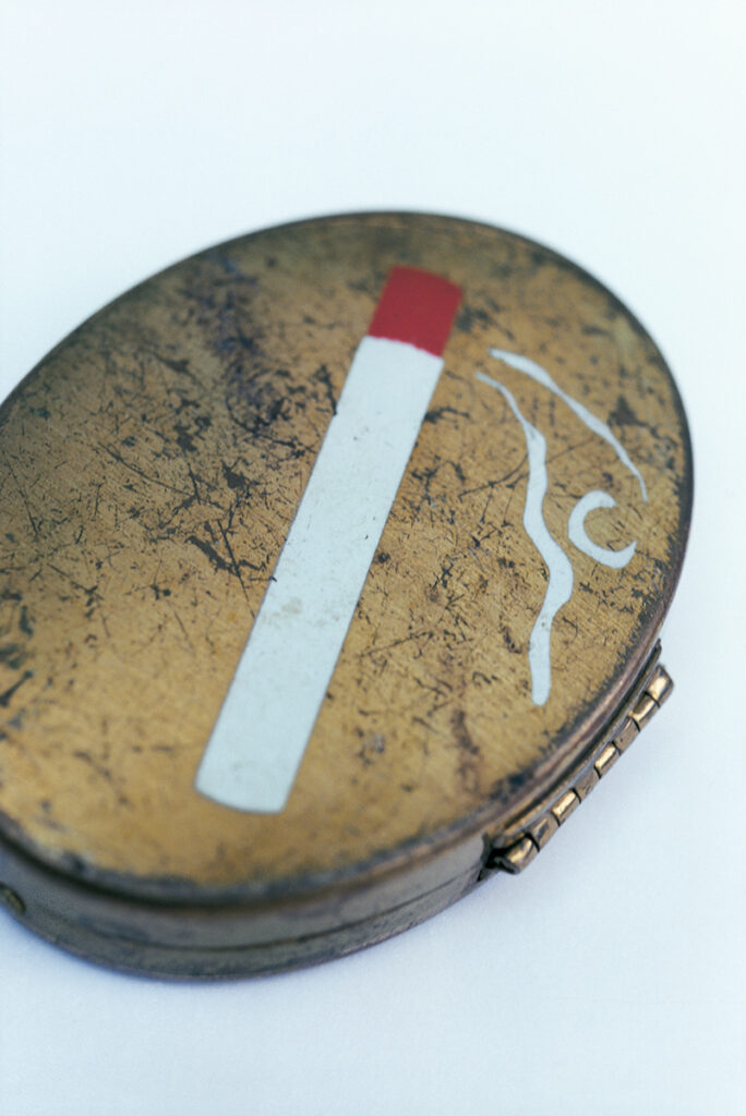 A close up image depicting an oval snuff box. There is a painting of a white cigarette with a red tip on the lid of the box.
