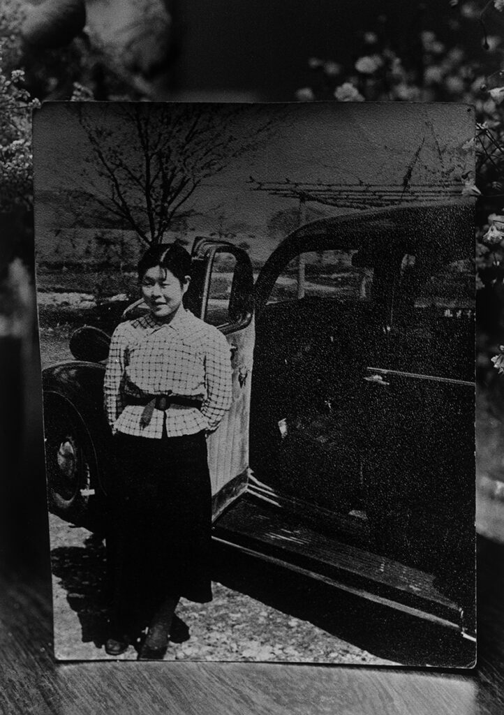 A photograph of a black and white photograph depicting a young woman standing next to a black cab. The door of the car is open, but the woman faces away from it, towards the camera.
