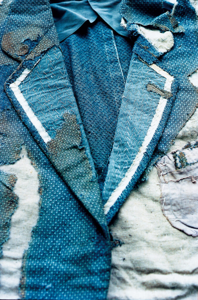 Close up of a blue collared jacket, comprising different fabrics in a patchwork. The garment is torn and damaged.