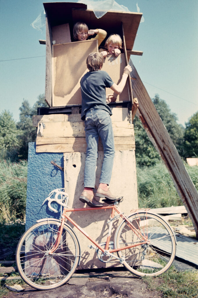 Colour photo. Two children peer from the window of a small, hand-built wooden tower. A third child looks in from the exterior, reaching the window by standing on the seat and frame of a red bicycle.