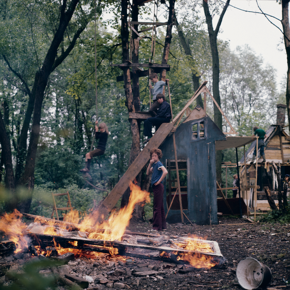 Colour image. Four children stand amid a makeshift structure resembling a house. A wooden pallet burns in the foreground.