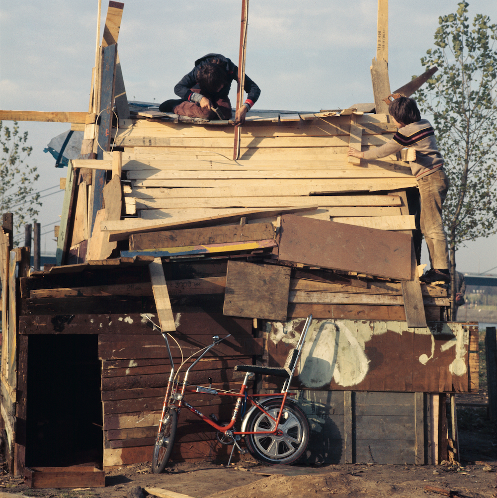 Colour photo. Two children are constructing the roof of a hand-built wooden hut. A red bicycle stands in the foreground.