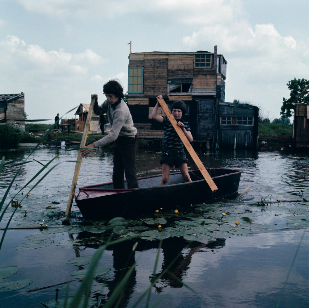 Colour image. Two children steer a small boat using wooden planks as oars. In the background we see a handmade wooden hut with windows.