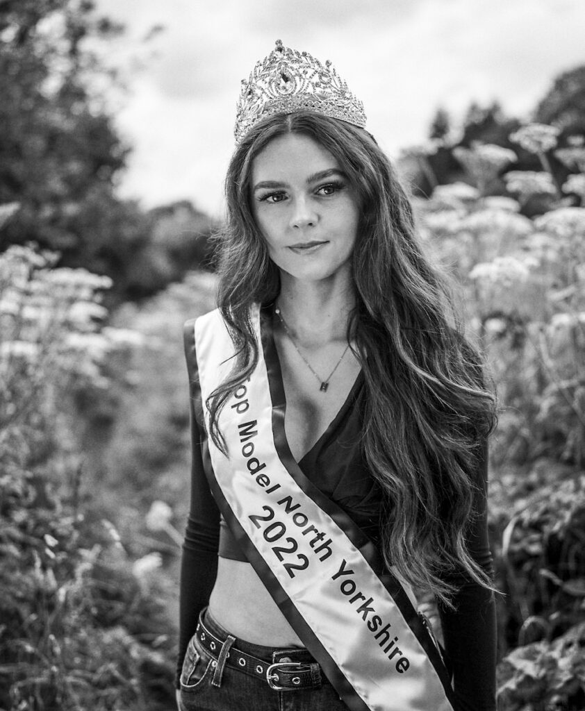 Black and white portrait of a young woman standing in a rural setting. She wears a tiara and a fabric sash bearing the words 'Top Model North Yorkshire 2022'.