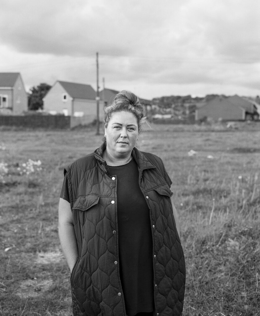 Black and white portrait of a woman standing in a rural setting. She wears a padded waistcoat and has her hair tied in a bun. She gazes directly at the camera.