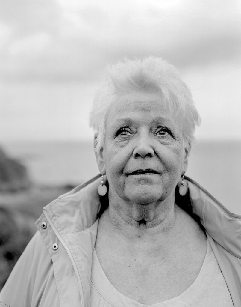 Black and white portrait of a woman in close up. She looks beyond the camera. The sky fills the background, with the horizon barely visible.
