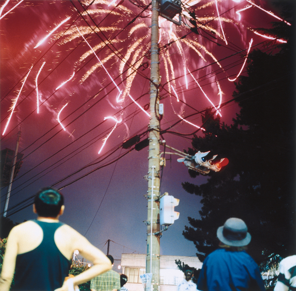 Colour photograph depicting a red firework bursting just beyond a telephone pole. A small crowd of people stands on the street below, observing it.