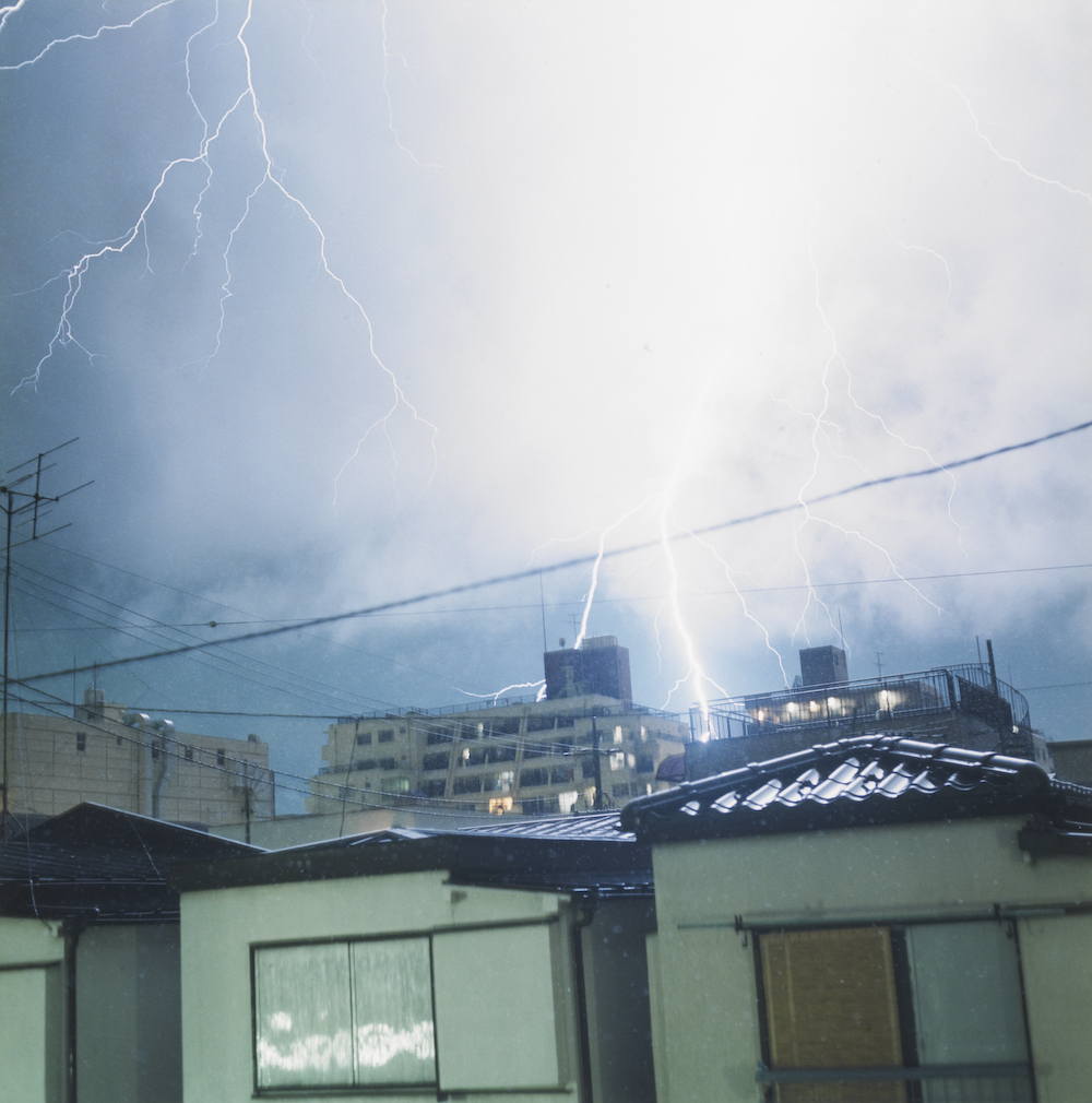 Colour photograph. A dark sky is lit with brilliant forks of lightning. In the foreground we see small, green-tinted houses, with taller buildings in the backround.