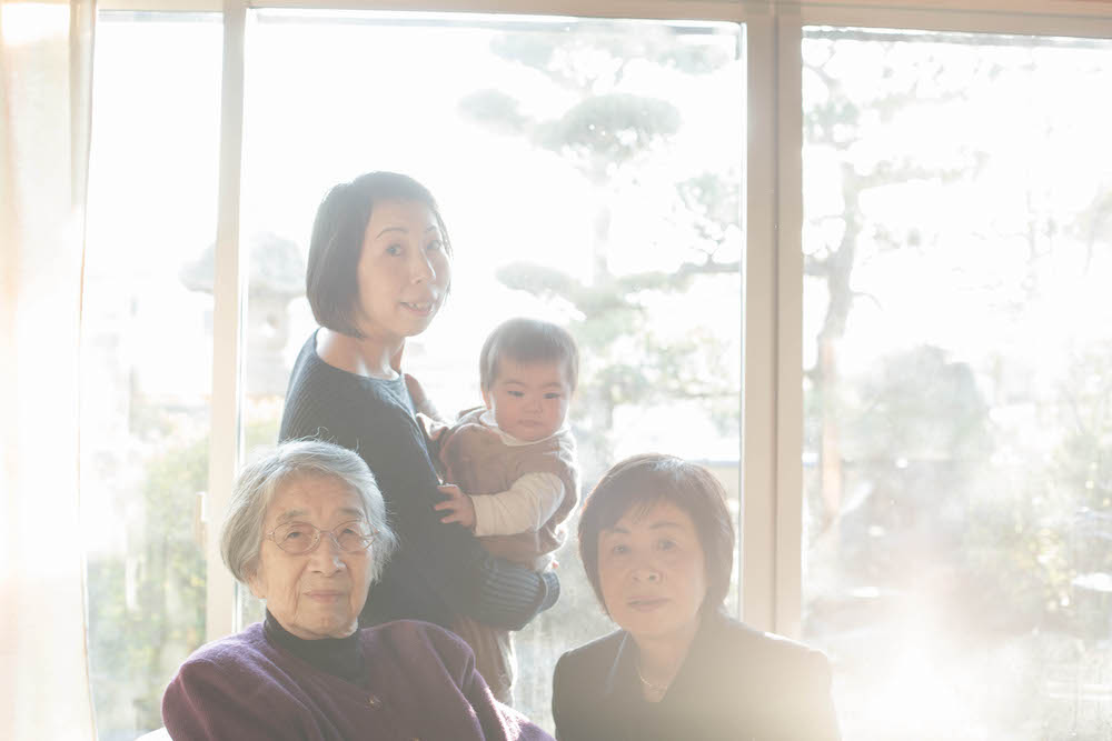 Colour photograph depicting four people - three women from different generations and a baby - sitting for a portrait in front of a window, bleached with sunlight.