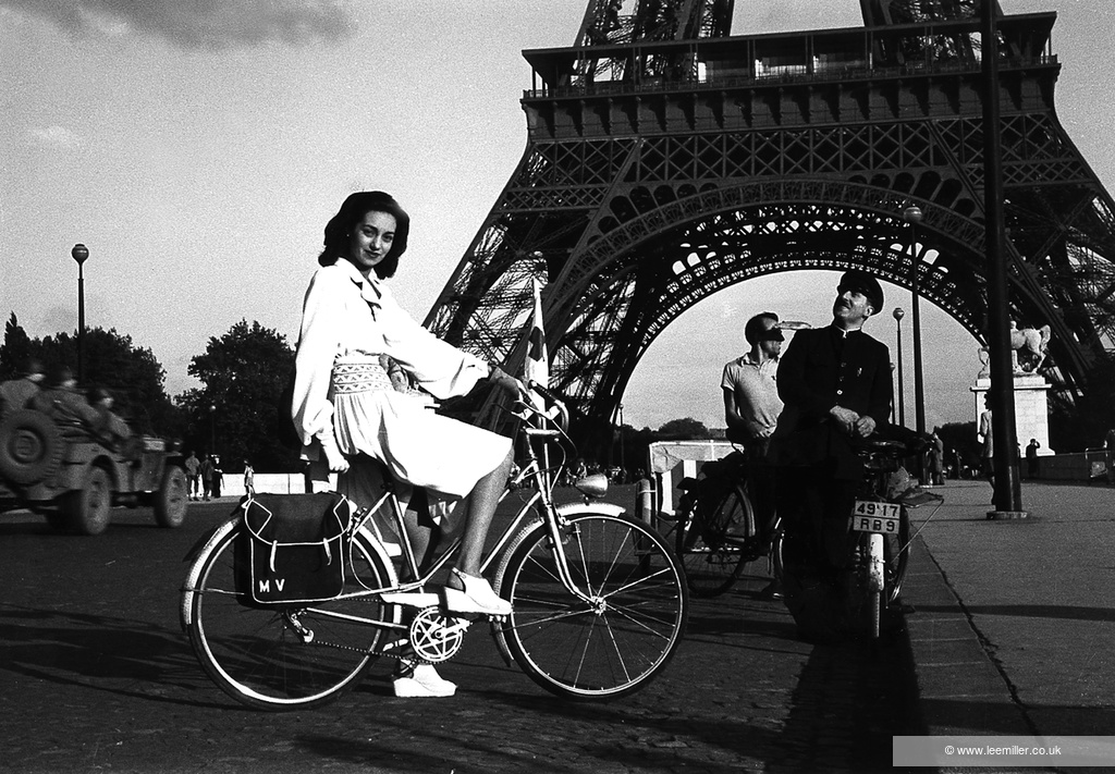 A young woman (Norma Wittig Cooke) sits atop a bicycle in front of the Eiffel Tower, Paris, while two men in the background look on. On the left, a military vehicle with soldiers drives past.