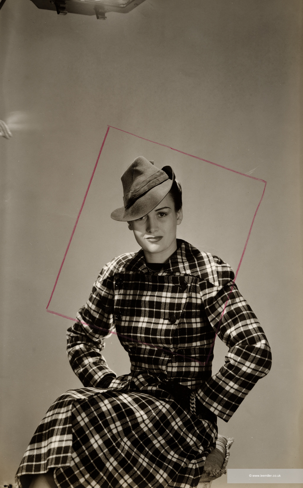 Sepia-toned photograph of a woman in a chequered jacket and soft hat, tilted slightly. A pink square, comprised of four thin lines, occupies the background.