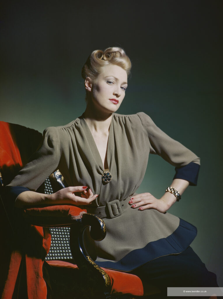 Colour photograph of a woman posing in an olive green jacket, secured with a blue and gold broach. She wears her blonde hair in curls, and leans against a red armchair.