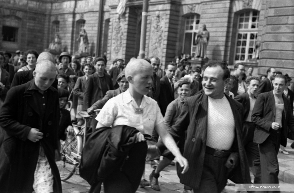 Black and white photograph. A crowd of people watch as women with shaved heads walk down a street.