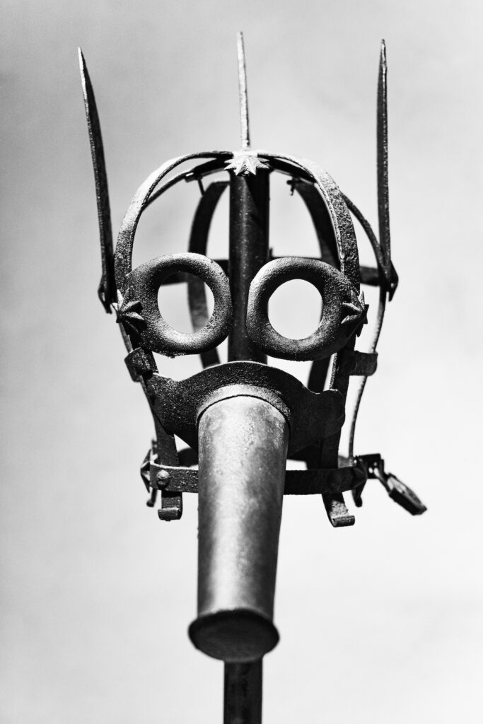 Black and white photograph of an iron cage mask, with a long mouthpiece, large eye holes, and vertical spikes protruding from the crown and sides..