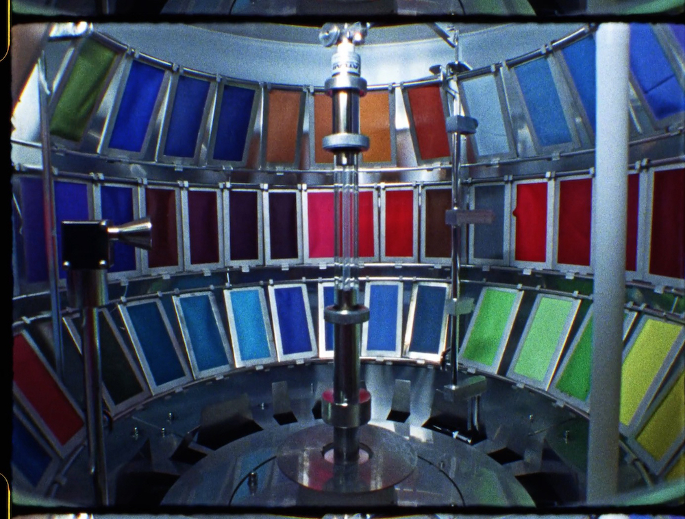 Film still depicting artificial dye being tested in a laboratory. Squares in a variety of colours are framed by metal apparatus.