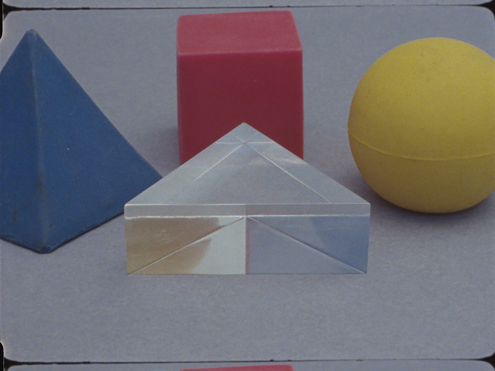 Film still depicting a yellow sphere, a red cube, and a blue triangular prism surrounding a transparent prism.