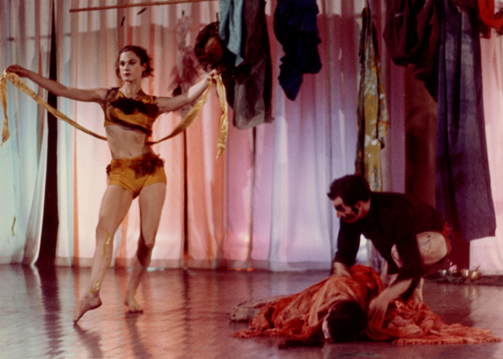 Photograph of a dance performance. One person lies on the floor, another crouches over them, while a woman stands at the left side of the image holding yellow streamers.