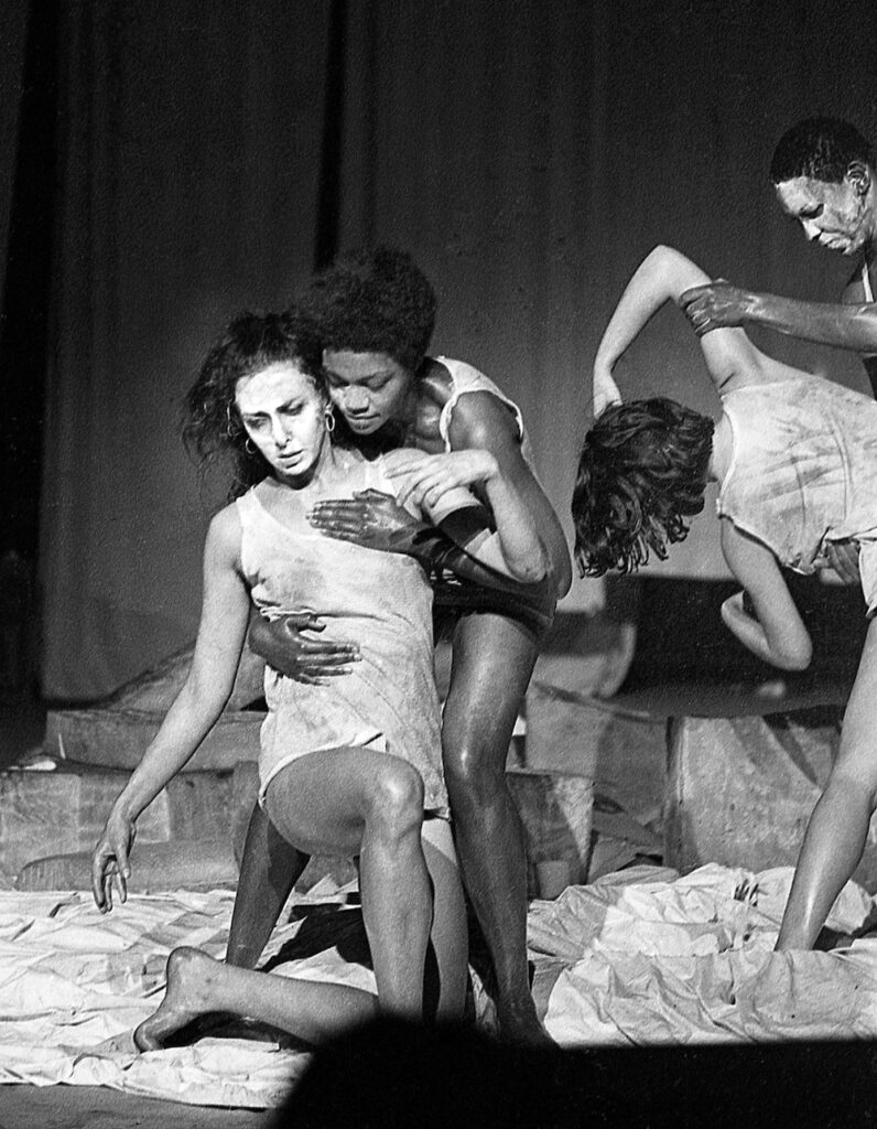 Black and white photograph. Two women, one of whom has painted her face white, embrace during a contemporary dance performance.