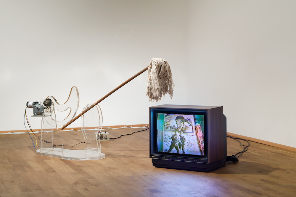 A motorised mop is suspended above a television. Both a sitting on a laminated floor, in front of a white wall.