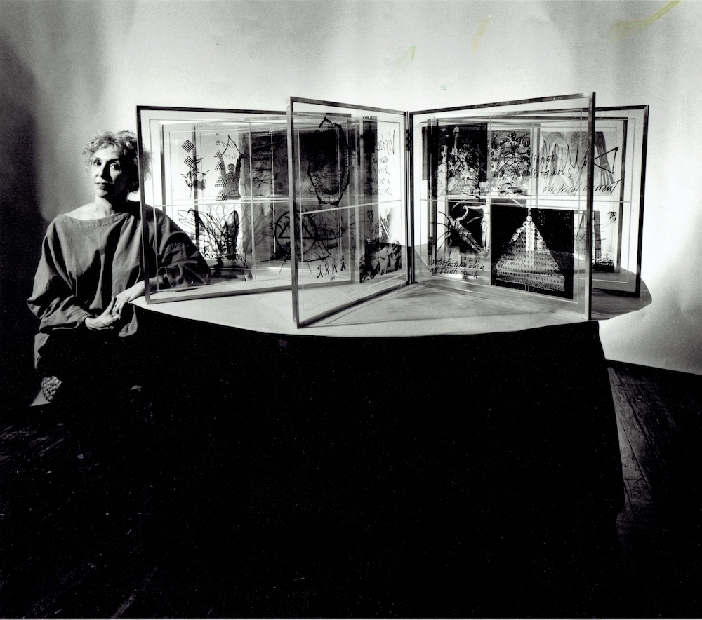 Black and white photograph. Carolee sits at the left on the image, next to a table displaying her work.