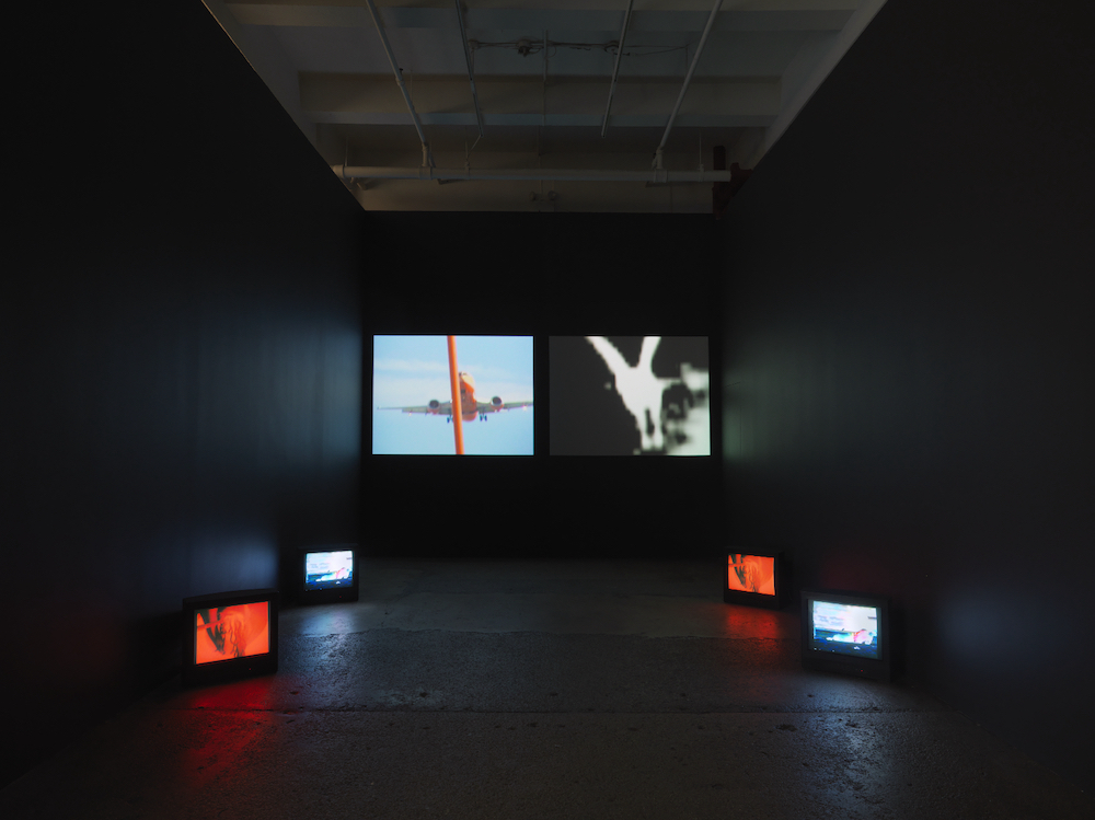 Installation image. Four televisions showing white and red images are arranged on the floor. Two larger screens comprise the back wall.