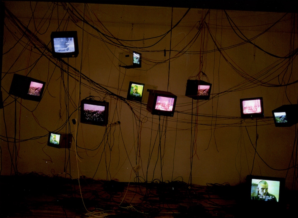 Colour photograph. Televisions are suspended from the ceiling, while one sits on the floor below. The screens are displaying a film.