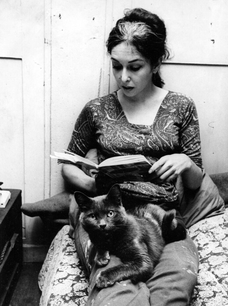 Black and white photograph. Carolee sits reading a book, her cat Kitch on her lap.