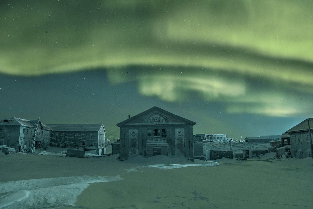 Colour photograph. A bright green aurora glimmers above a group of buildings. The landscape is entrenched in snow.