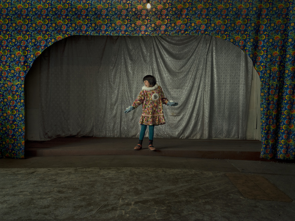 Colour photograph. A young girl in a flower patterned dress standing beneath an archway, also patterned with flowers. A silver curtain hangs in the background.
