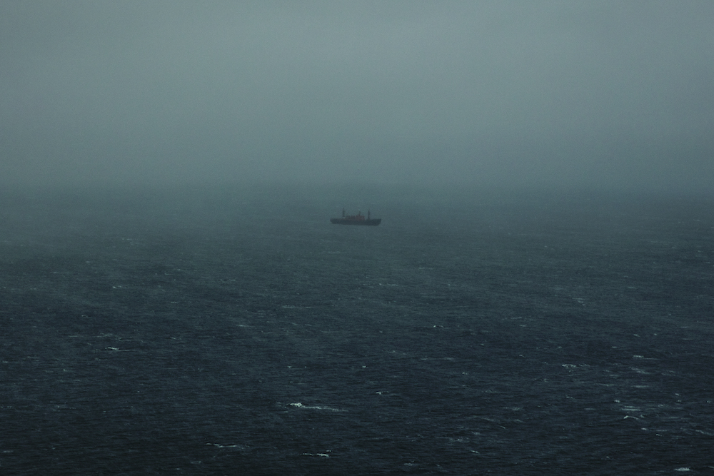 Colour photograph from Hyperborea. A small boat in the distance, surrounded by a great expanse of sea. It is misty.