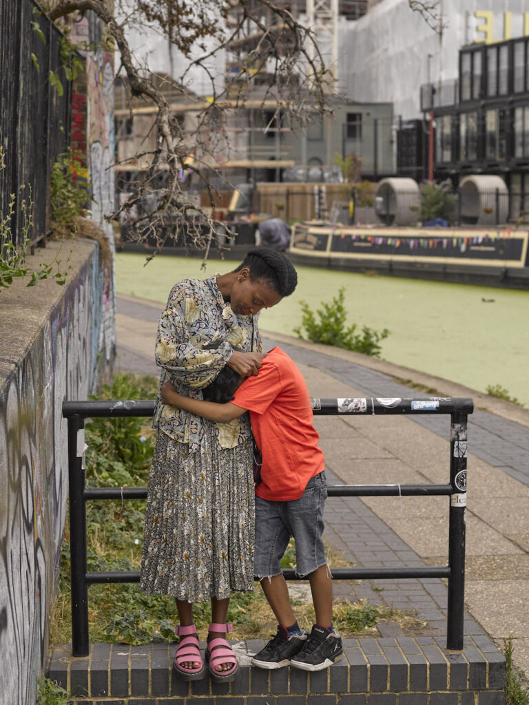 Colour photograph. A woman in a patterned grey skirt and shirt embraces a young boy in an orange t-shirt and shorts. They are standing on a small wall, in front of a black railing. There is a canal in the background, with a canal boat decorated in colourful bunting.