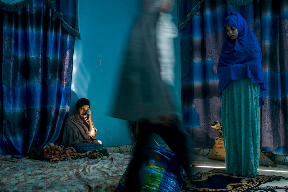 Colour photograph, comprising a palette of blue tones. One woman sits on a blanket at the left side of the frame, with a woman stood at the right wearing a blue veil and dress. A third figure passes through the centre of the image, a blur of motion.
