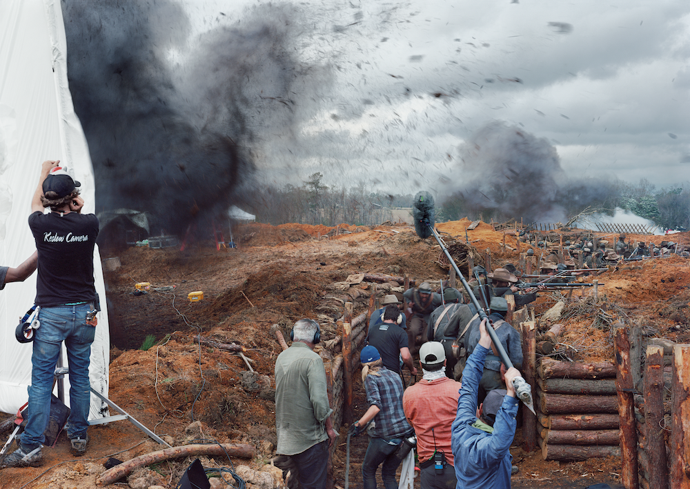 Colour photograph of a film set. A film crew is assembled in the foreground, with soldiers in trenches towards the rear. A boom pole stretched across the image, capturing the sound of a simulated explosion. Shrapnel flies through the air.