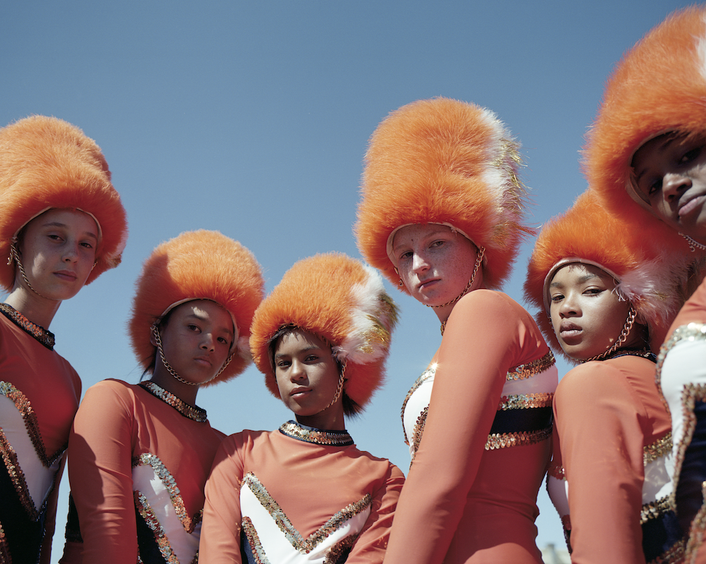 Colour photograph. Six young women in band uniform (large orange hats and sequined clothes) gaze down the lens.
