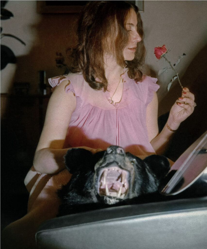 Colour photograph. A woman in a pink blouse is shown in profile, her face turned towards the red rose she is holding. Slightly out of focus in the nearer foreground is a taxidermied black bear.