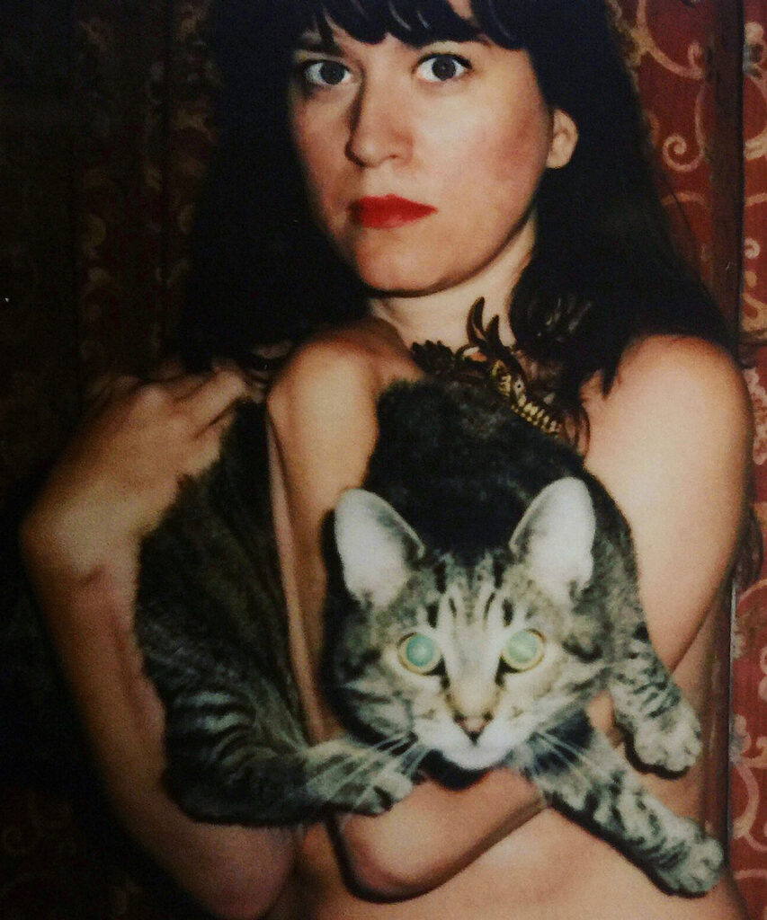 Colour photograph. A woman wearing red lipstick is holding a tortoiseshell cat. Both look directly into the lens.