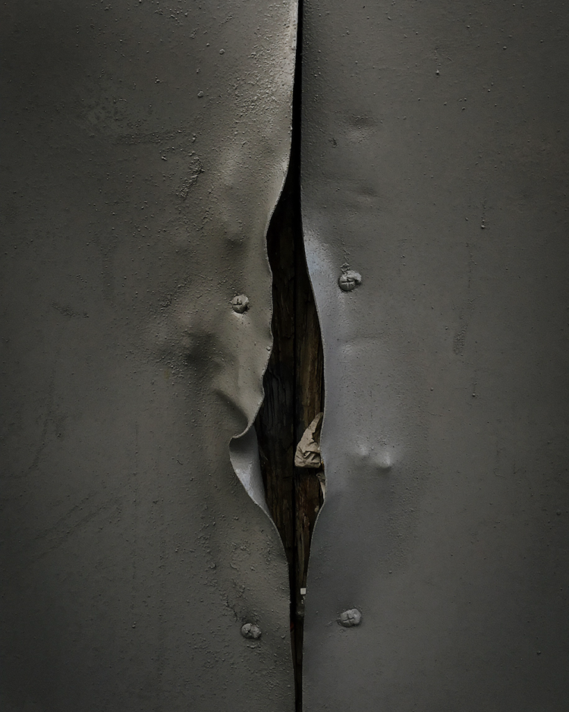 Colour photograph. Two sheets of grey metal are coming apart, revealing a dark fissure.