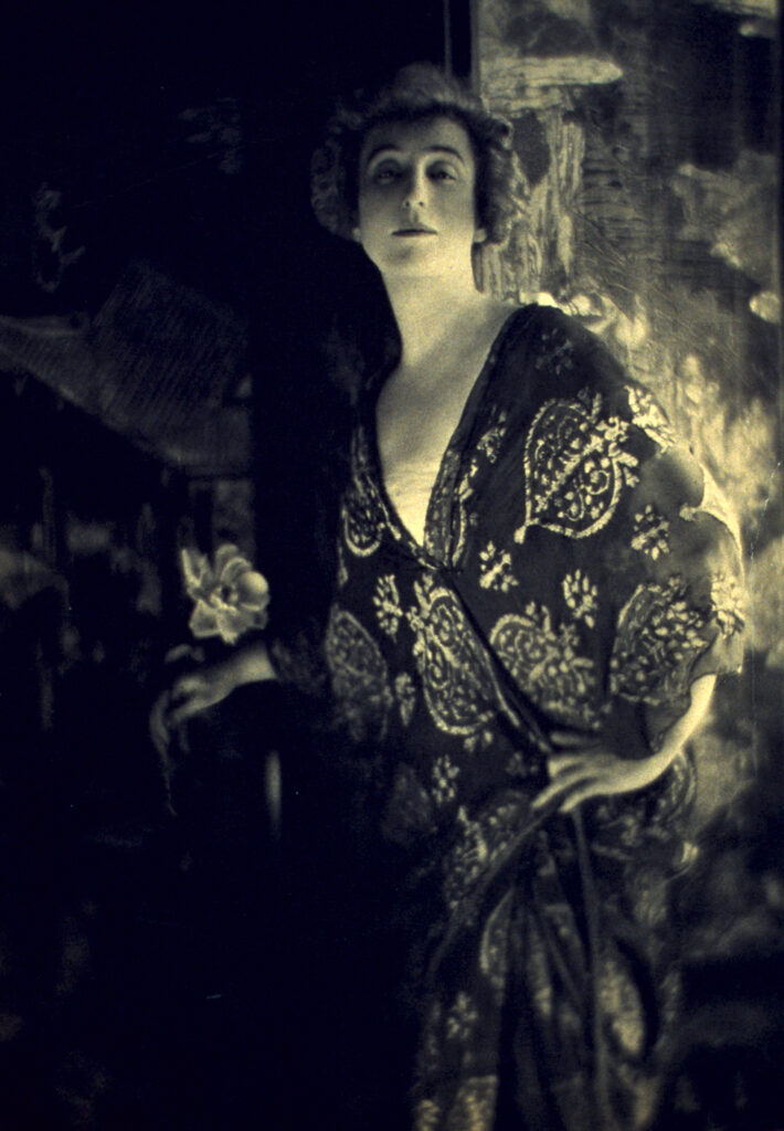 Slightly yellow toned, black and white photograph. A woman (Ava Lowle Willing) poses in a patterned robe, her hand on her hip.