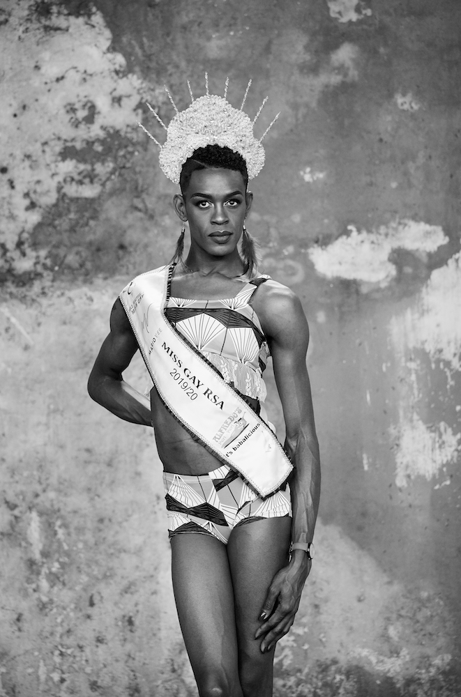 Black and white photograph. The subject poses in a tiara and a sash which reads 'Miss Gay RSA 2019/20'.