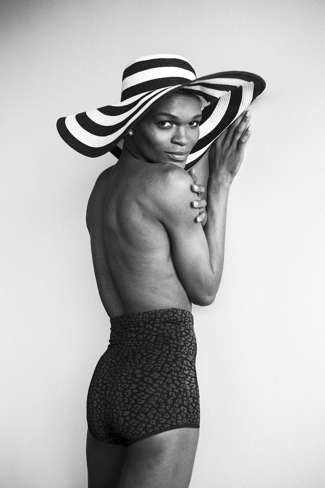 Black and white photograph. Portrait of a person wearing a large, wide brimmed, stripy sun hat and animal print shorts.