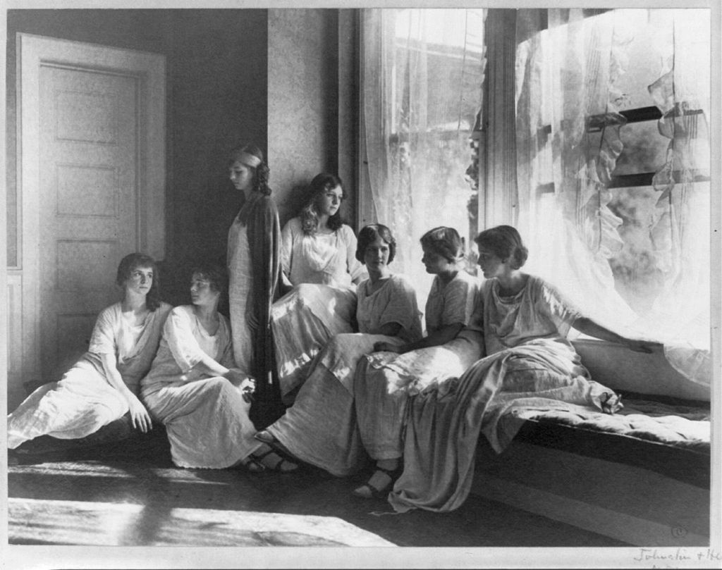 Black and white photograph of a group of women in white dresses. They are sitting / standing in front of a large window, through which sunlight is streaming.