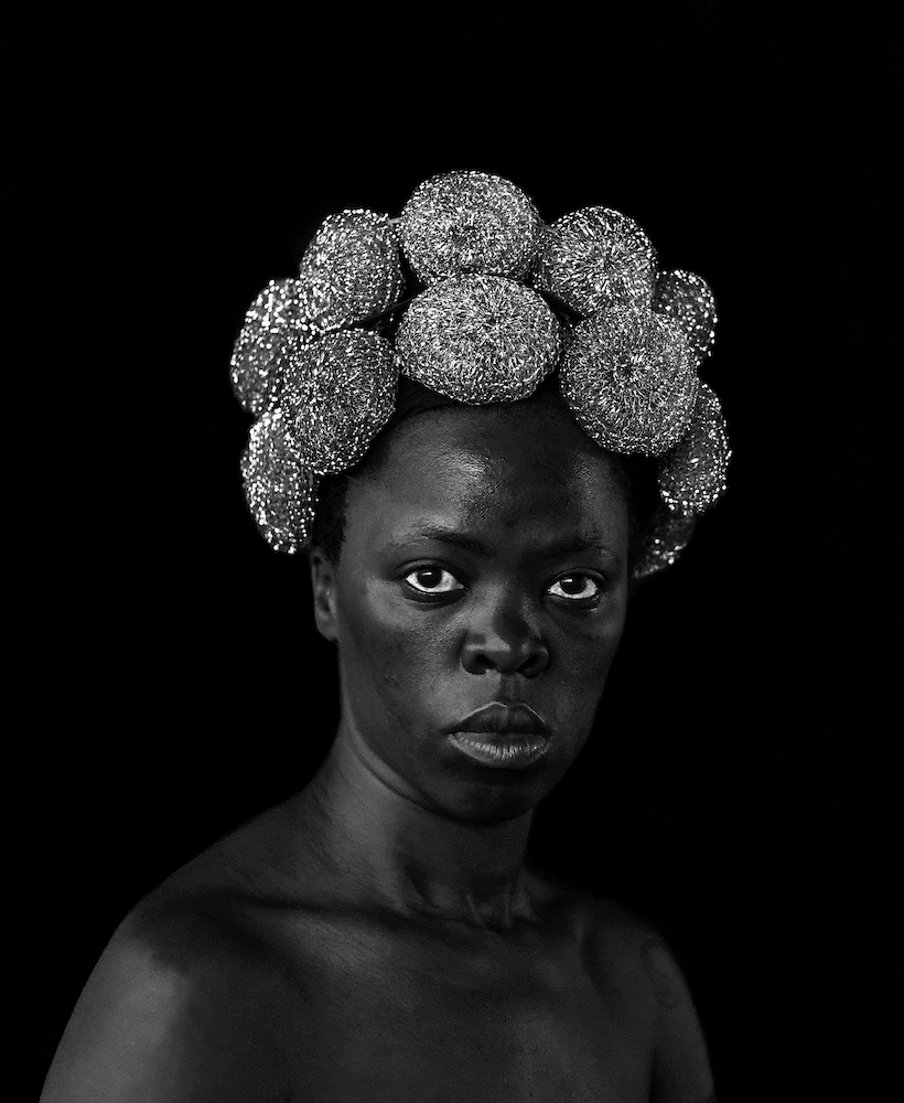 Black and white photograph. Zanele looks towards the camera, their hair decorated with scouring pads.