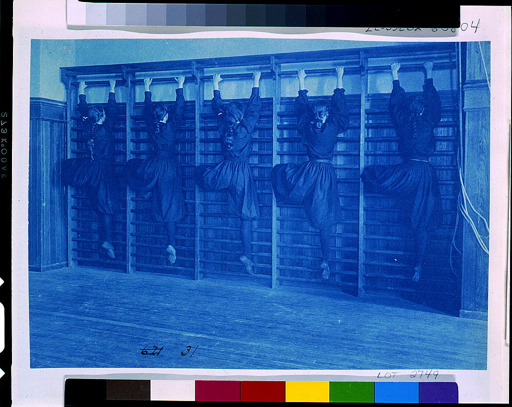 Cyanotype, blue toned photograph. Five girls are faced away from the camera, climbing wooden climbing apparatus in a school gym hall.